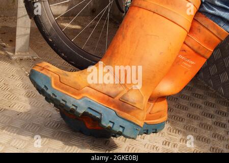 Vue rapprochée des pieds d'un homme portant des bottes de pêche orange et bleu sur le pont métallique d'un bateau devant une roue à vélo Banque D'Images