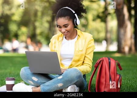 Une étudiante africaine, une fille qui s'est apprenante sur un ordinateur portable, porte des écouteurs dans Park Banque D'Images