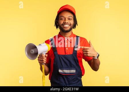 Bon homme de travail à barbe portant une combinaison bleue et un T-shirt rouge regardant la caméra, tenant le mégaphone entre les mains, annonçant la publicité. Intérieur s Banque D'Images