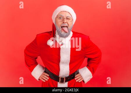 Désobéissant drôle homme âgé avec barbe grise portant le costume du père noël démontrant la langue, se comportant méchant indiscipliné, humeur enfantine. Studio intérieur Banque D'Images