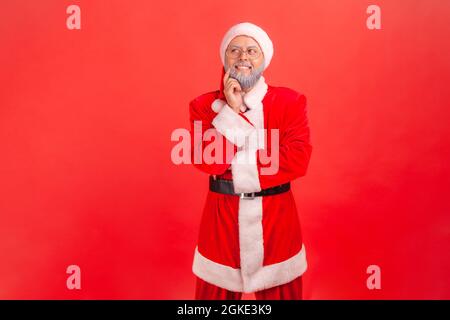 Homme âgé rêveur avec barbe grise portant le costume du père noël debout avec le doigt sur la joue rêvant et pensant à des souvenirs agréables. Studio intérieur Banque D'Images