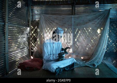 Ahmed sur un tapis de raphia au sol d'une cabane jouant avec deux oiseaux, portant les vêtements blancs d'un imam, Mongla, Sundarbans, Bangladesh Banque D'Images