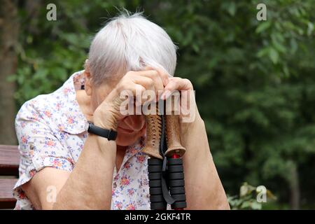 Femme âgée assise avec sa tête abaissée sur des bâtons de marche sur un banc. Maladie et fatigue, mode de vie sain à la vieillesse, vie à la retraite Banque D'Images