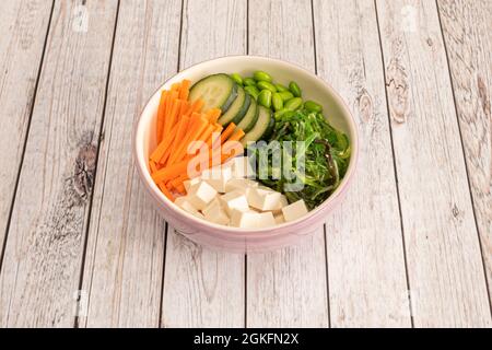 Bol vegan rose avec salade d'algues wakame et tofu blanc coupé en dés, carottes râpées, tranches de concombre et haricots édamames sur table en bois Banque D'Images