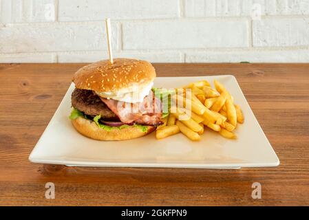 Hamburger de bœuf avec bacon, oignon rouge, laitue iceberg et œuf frit garni de frites sur une assiette carrée blanche Banque D'Images