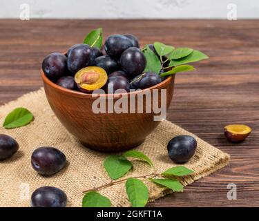 Prunes fraîches mûres entières et tranchées avec des feuilles dans un bol sur des tapis de toile sur une table en bois brun.Récolte d'été. Banque D'Images