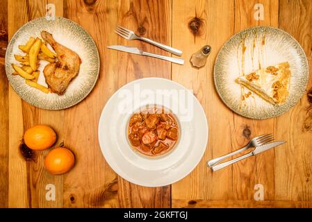 Menu pour une personne avec ragoût de viande, cuisse de poulet rôti avec pommes de terre de luxe, tarte à la crème avec noix. Couteaux et fourchettes sur une table en pin Banque D'Images