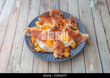 Assiette bleue pleine d'œufs frits au jambon de serrano, tranches de pommes de terre avec paprika sur une table en bois Banque D'Images