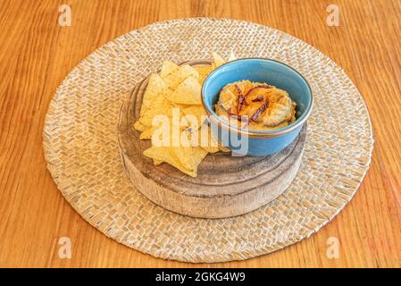 Petit bol bleu avec houmous de pois chiches, flocons de poivre séché et nachos à tremper sur un morceau de log et un trivet en osier Banque D'Images