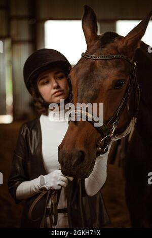 Belle fille jockey, à côté de gros plan de tir d'un beau cheval brun. Une belle jeune fille se tient à côté d'un cheval dans l'écurie. Ranch. Équitation. Hippodrome Banque D'Images