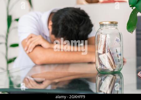 jeune homme crevé avec ses économies dans un pot en verre sur la table, billets à l'intérieur d'un pot en verre. garçon inquiet et s'ennuiait avec ses économies et Banque D'Images