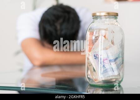 jeune homme crevé avec ses économies dans un pot en verre sur la table, billets à l'intérieur d'un pot en verre. garçon inquiet et s'ennuiait avec ses économies et Banque D'Images