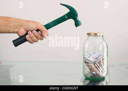 homme avec un marteau dans une banque de pigeons brisant la main. pot en verre avec des billets, des économies et des finances personnelles. Fond blanc. Concept d'économie Banque D'Images