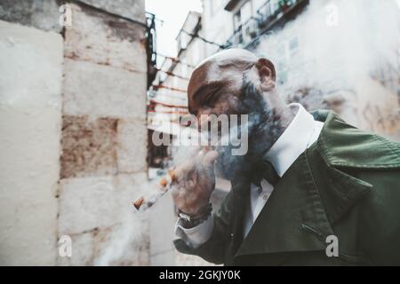 Une vue latérale d'un élégant homme noir à barbe mûr dans une tenue à la mode vert foncé fumant un cigare à l'extérieur ; un portrait d'un élégant chauve-Africain b Banque D'Images