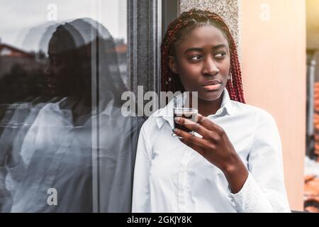 Portrait d'une jeune femme noire éblouissante avec des tresses rouges et dans une chemise blanche, penchée contre un mur de verre tout en tenant dans la main une tasse de morni Banque D'Images