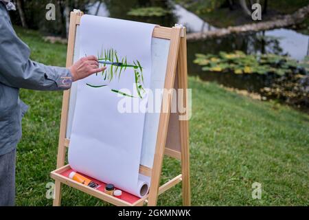 Une femme heureuse tire sur le chevalet avec une brosse et des peintures. Femme artiste dessine la nature et les arbres sur papier au bord de l'eau sur la rive de la rivière Banque D'Images