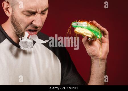 Le concept de restauration rapide et d'écologie. Un homme chauve avec une barbe, tenant un hamburger et cracher plastique de remplissage. Arrière-plan Bourgogne. Gros plan. Banque D'Images