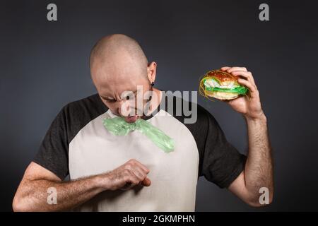 Le concept de restauration rapide et d'écologie. Un homme chauve avec une barbe, cracher des ordures remplissant un hamburger, étouffant. Arrière-plan sombre. Copier l'espace. Banque D'Images