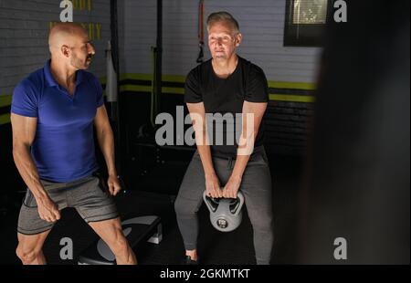 Entraîneur personnel masculin aidant l'homme mature à faire l'exercice Kettlebell Swing dans la salle de gym à la maison Banque D'Images