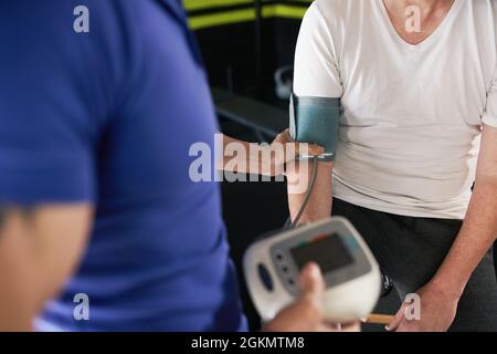 Gros plan de la santé masculine et de bien-être entraîneur personnel aidant l'homme âgé senior dans la mesure de la tension artérielle avant l'entraînement dans la salle de gym à la maison petit buline Banque D'Images