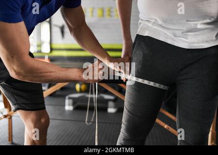 Gros plan de deux personnes méconnues. Entraîneur personnel mesurant les clients cuisse avec mètre ruban dans la salle de gym à la maison Banque D'Images