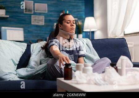 Femme avec col cervical assis sur un canapé souffrant de douleur de colonne vertébrale. Jeune caucasien avec une blessure physique prenant un médicament contre la douleur après un accident du dos, avec une gêne Banque D'Images