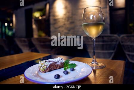 Des cannoli siciliens roulent avec des myrtilles sur une plaque de céramique blanche et un verre de vin blanc sur une table de restaurant avec un décor de restaurant dans le backgro Banque D'Images