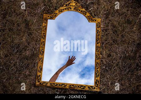 Miroir doré vintage couché sur fond sombre reflétant la main mystérieuse qui s'atteint sur le ciel bleu et les nuages. Vue de dessus. Main d'aide Banque D'Images