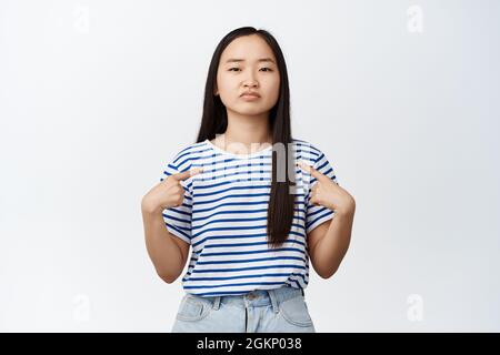 Image de triste brune asiatique fille pointant les doigts sur elle-même, faisant le visage bouleversé de l'expression, debout dans un tshirt rayé sur fond blanc Banque D'Images