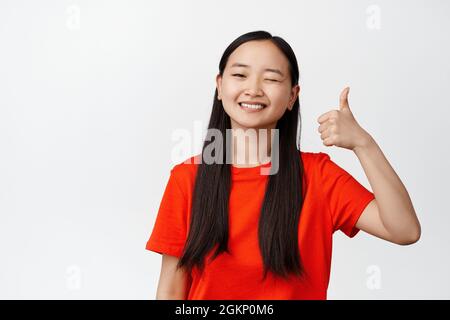 Souriante fille coréenne montre des pouces et des wencing, des commentaires positifs, comme et d'approuver quelque chose de bon, se tenant sur fond blanc Banque D'Images