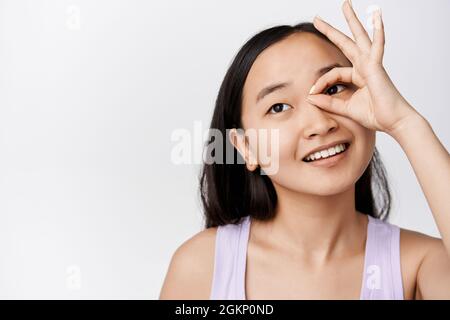 Beauté. Jeune femme asiatique avec une peau claire et brillante, montrant ok, aucun geste contre les yeux, souriant et regardant loin, debout sur fond blanc Banque D'Images