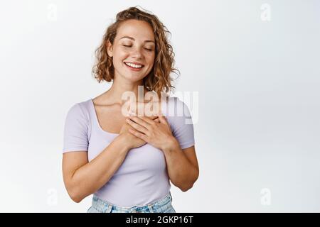 Bonne femme blonde tenant les mains sur le coeur et souriant, les yeux proches réfléchis, rêvant de sourire romantique, rêvant de jour, debout sur fond blanc Banque D'Images