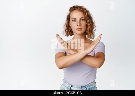 Une jeune femme sérieuse montre un geste d'arrêt de croix, regardant sans amused, disant non, interdit quelque chose, se tenant sur fond blanc Banque D'Images