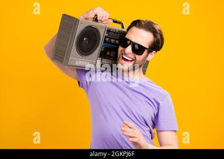 Photo de l'excitée cool jeune gentleman porter un t-shirt violet lunettes foncé sourire écoute boîte de perche isolée couleur jaune fond Banque D'Images