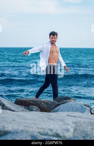 Vue latérale d'un homme élégant et serein en chemise blanche marchant le long de la côte rocheuse sur fond de mer bleue et regardant l'appareil photo Banque D'Images