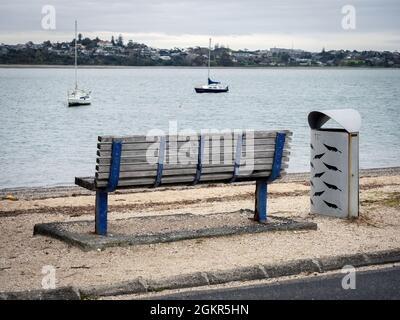 AUCKLAND, NOUVELLE-ZÉLANDE - 07 juillet 2021 : banc en bois solitaire à Bucklands Beach à Auckland, Nouvelle-Zélande avec de petits bateaux en arrière-plan Banque D'Images