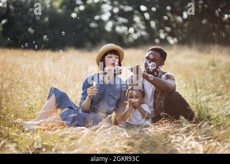 Joyeuse race mixte famille de trois assis ensemble sur l'herbe et bulles de savon. Des parents heureux jouant avec un petit fils mignon sur la nature. Pique-nique et plaisir. Banque D'Images