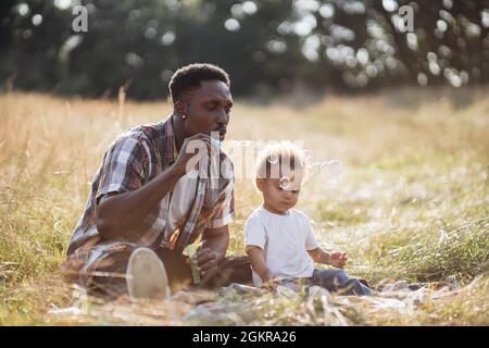 Afro-américain homme dans des vêtements décontractés assis avec un petit garçon sur l'herbe et bulles de savon soufflantes. Papa heureux jouant avec son mignon sur l'air frais. Concept de la famille et de l'heure d'été. Banque D'Images