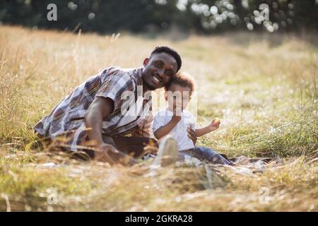 Père afro-américain embrassant son petit fils et souriant à la caméra tout en étant assis ensemble sur le terrain d'été. Un adorable garçon mangeant du raisin doux pendant le pique-nique avec un père attentionné. Banque D'Images