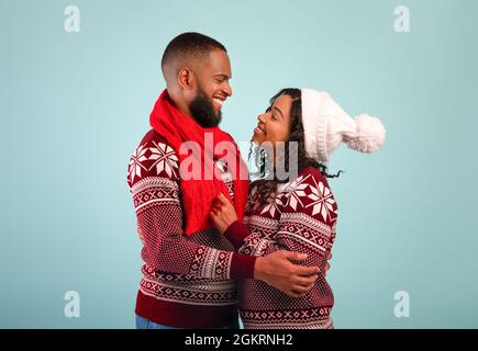 C'est Noël. Couple afro-américain aimant dans des sweaters chauds embrassant et regardant l'un l'autre, fond bleu Banque D'Images