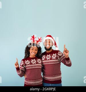 Offre incroyable pour Noël. Un couple afro-américain enthousiaste dans des chandails pointant vers le haut de l'espace libre, fond bleu Banque D'Images