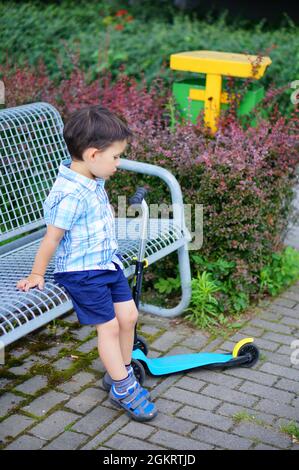 POZNAN, POLOGNE - 15 juillet 2017 : un garçon assis sur un banc de métal à côté de son scooter dans un parc Banque D'Images