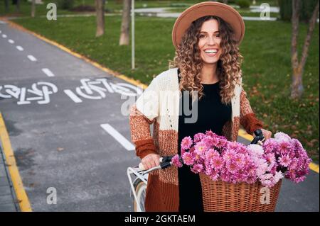 Jolie fille gaie avec un doux sourire à cheval sur vélo avec bouquet de fleurs dans le panier. En arrière-plan, pistes cyclables et pelouses vertes. Banque D'Images