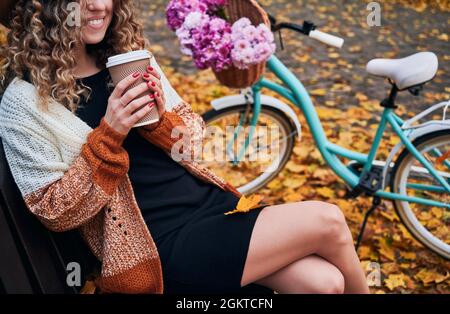 Jeune femme souriante assise sur le banc près du vélo avec des fleurs et de boire du café pour aller dans le parc d'automne. Femme élégante tenant une tasse de boisson chaude et souriante tout en se reposant à l'extérieur près de la ville vélo. Banque D'Images