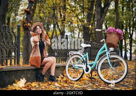 Une femme positive assise sur une bordure en béton de la clôture du parc près de son vélo de ville bleu vif à la chaude journée d'automne. Activités sportives, balade en vélo dans les espaces ouverts de la ville. Banque D'Images