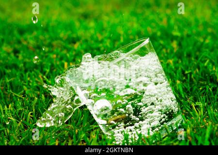 Chute d'un verre d'eau ou de limonade sur fond d'herbe. L'eau avec des éclaboussures et des gouttes coule d'un verre Banque D'Images