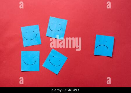 Un triste visage regardant des visages souriants de loin. Visages heureux et tristes dessinés sur des autocollants bleus. Concepts d'exclusion, de différence, de jalousie. Banque D'Images