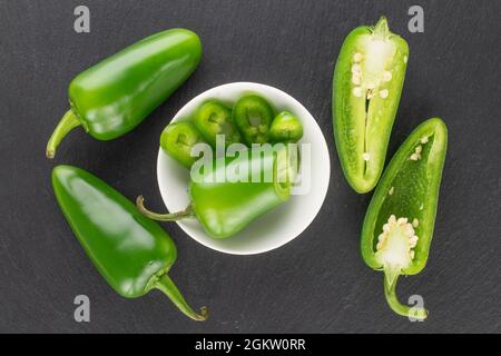 Deux gousses entières de poivrons verts chauds et coupées en tranches dans une soucoupe blanche sur une pierre d'ardoise, vue rapprochée du dessus. Banque D'Images