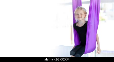 Banner enfants exercices de yoga aérien. Portrait jeune fille souriante s'entraîner dans l'oscillation d'étirement aéro dans hamac pourpre dans le club de fitness. CopySpace Banque D'Images