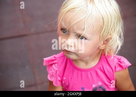 Grave fille de bébé en colère regardant à gauche. Blonde blu oeil enfant dans le portrait de t-shirt rose. Revêtement en caoutchouc protecteur flou du terrain de jeu en arrière-plan Banque D'Images